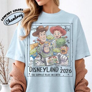 May include: Light blue t-shirt featuring a graphic of Toy Story characters, including Woody, Buzz Lightyear, and Jessie. The design includes the text "DISNEYLAND 2026" and "THE HAPPIEST PLACE ON EARTH". The shirt is labeled "COMFORT COLORS Chambray".
