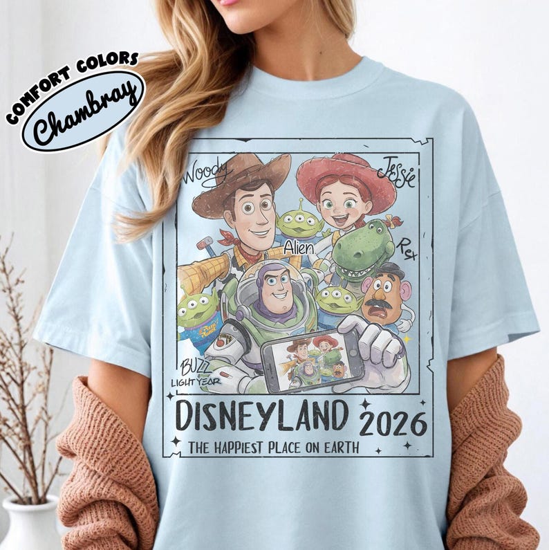 May include: Light blue t-shirt featuring a graphic of Toy Story characters, including Woody, Buzz Lightyear, and Jessie. The design includes the text "DISNEYLAND 2026" and "THE HAPPIEST PLACE ON EARTH". The shirt is labeled "COMFORT COLORS Chambray".