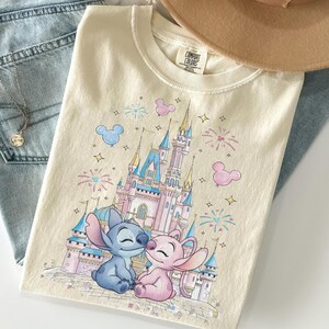 May include: A cream-colored t-shirt featuring a graphic print of a blue alien character and a pink alien character sitting in front of a large castle. The design includes colorful fireworks, star shapes, and Mickey Mouse-shaped balloons.