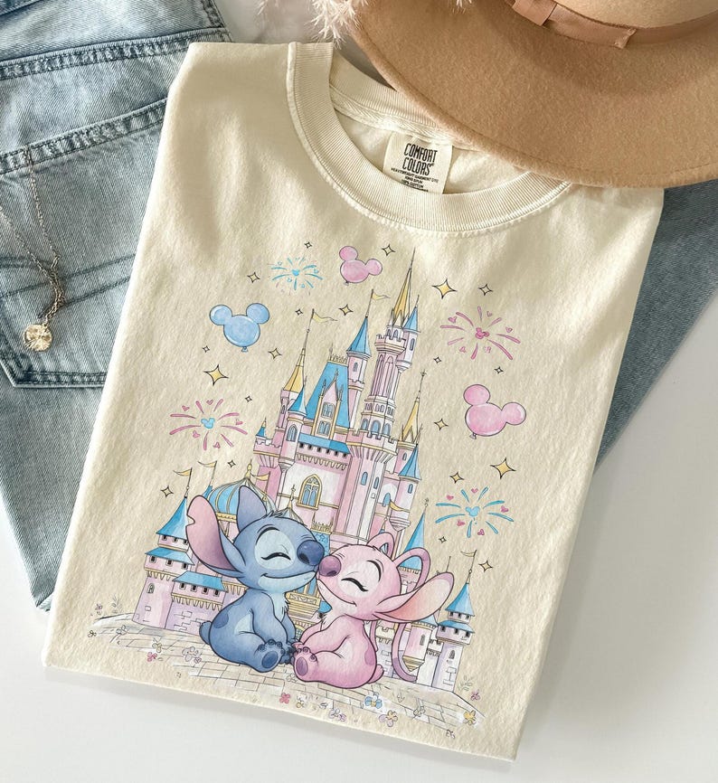 May include: A cream-colored t-shirt featuring a graphic print of a blue alien character and a pink alien character sitting in front of a large castle. The design includes colorful fireworks, star shapes, and Mickey Mouse-shaped balloons.