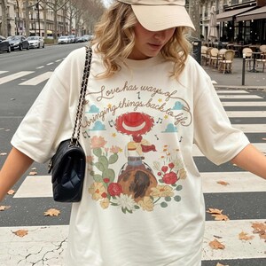 May include: An off-white short-sleeve t-shirt featuring a graphic of a cowgirl on a horse surrounded by cacti and flowers. The text reads, Love has ways of bringing things back to life. A black quilted shoulder bag with a chain strap is also visible.