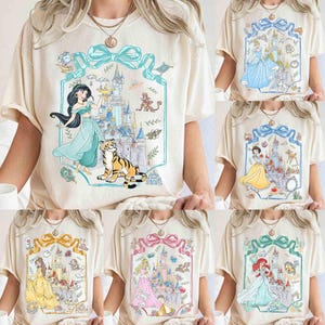 K&ouml;nnte beinhalten: Cremefarbene T-Shirts mit verschiedenen Disney-Prinzessinnen-Designs. Jedes Shirt zeigt eine Prinzessin mit einem Schloss im Hintergrund, umrahmt von einem dekorativen Rahmen mit einer Schleife. Die Prinzessinnen sind Jasmin, Aschenputtel, Schneewittchen, Belle, Aurora und Arielle.