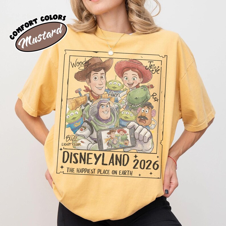 May include: Mustard-colored t-shirt featuring a graphic of Disney's Toy Story characters, including Woody, Buzz Lightyear, and Jessie. The design includes the text "DISNEYLAND 2026" and "THE HAPPIEST PLACE ON EARTH". The shirt is a Comfort Colors brand.