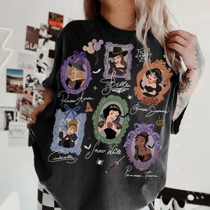 May include: Dark gray t-shirt featuring framed portraits of Disney princesses, including Aurora, Belle, Jasmine, Cinderella, Snow White, and Tiana. The shirt has a vintage, distressed look with the princesses' names in cursive.