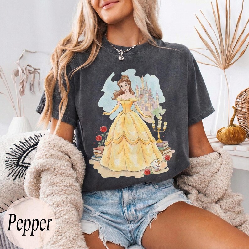 K&ouml;nnte beinhalten: Dunkelgraues T-Shirt mit einer farbenfrohen Illustration einer Prinzessin in einem gelben Kleid, einem Schloss und dem Text "Pepper". Das Design umfasst Rosen, B&uuml;cher und eine Teetasse. Das Shirt ist aus weichem Material.