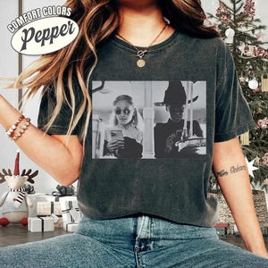 May include: Dark gray Comfort Colors Pepper t-shirt featuring a black and white image of two people. One is wearing sunglasses and holding a phone, the other is wearing a hat. The shirt has a distressed look.
