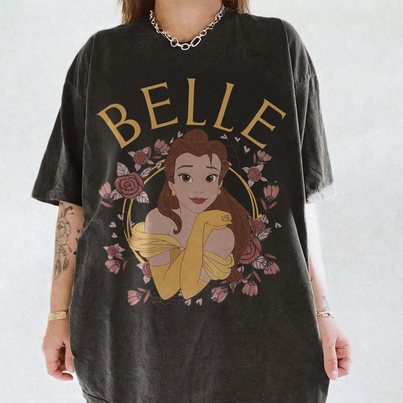 May include: A charcoal gray short-sleeve t-shirt featuring a graphic print of the character Belle from Beauty and the Beast. The design includes the text BELLE in gold letters above the character, surrounded by a circular frame of pink roses.