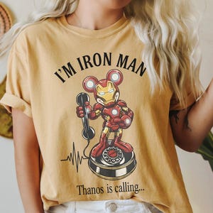 Op de afbeelding: Mosterdgeel T-shirt met de tekst "I'M IRON MAN" boven een cartoon Iron Man-personage met Mickey Mouse-oren die een telefoon vasthoudt. De zin "Thanos is calling..." staat eronder gedrukt.