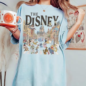 May include: A light blue t-shirt featuring a large graphic print with the text THE DISNEY above a detailed illustration of a castle and various animated characters. The shirt has short sleeves and a crew neckline, showcasing a colorful scene.