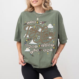 May include: An olive green short-sleeve t-shirt featuring a graphic print with the text "ITS A Toy Story BABY JUST YES SAY". The design includes rope-style lettering, colorful beads, white clouds, musical notes, and yellow star accents.