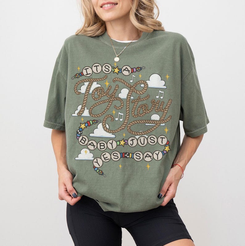 May include: An olive green short-sleeve t-shirt featuring a graphic print with the text "ITS A Toy Story BABY JUST YES SAY". The design includes rope-style lettering, colorful beads, white clouds, musical notes, and yellow star accents.