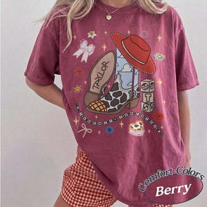 May include: A berry-colored short-sleeve t-shirt featuring a graphic print of a cowboy boot with a cow print pattern, a red cowboy hat, a bow, and blocks with the letters T, S, and 13. The text TAYLOR and I KNEW YOU are printed on the shirt.