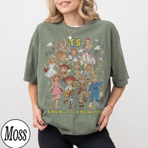 K&ouml;nnte beinhalten: Ein olivgr&uuml;nes Kurzarm-T-Shirt mit einem Grafikdruck von mehreren Zeichentrickfiguren in verschiedenen Outfits. Der Text "TS" erscheint oben und der Text "I KNEW IT, I KNEW YOU" ist am unteren Rand des Grafikdesigns aufgedruckt.