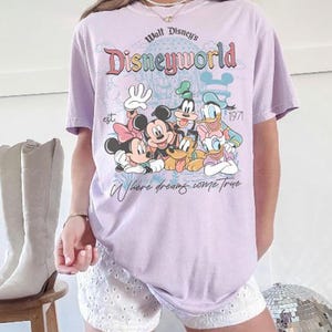 Puede incluir: Camiseta lila claro con la inscripci&oacute;n "Walt Disney's Disneyworld" y un dibujo de Mickey, Minnie, Goofy, Donald y Pluto. La camiseta tambi&eacute;n incluye el texto "est. 1971" y "Where dreams come true."