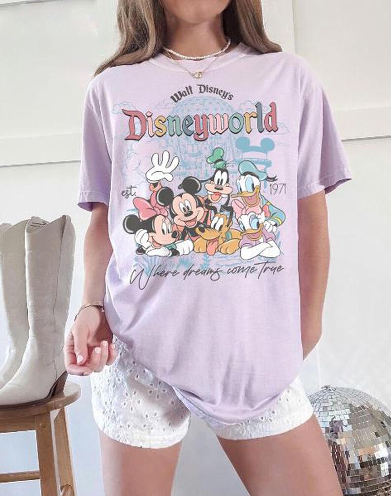 Puede incluir: Camiseta lila claro con la inscripci&oacute;n "Walt Disney's Disneyworld" y un dibujo de Mickey, Minnie, Goofy, Donald y Pluto. La camiseta tambi&eacute;n incluye el texto "est. 1971" y "Where dreams come true."
