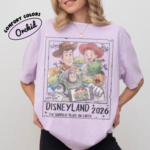 May include: A light purple t-shirt featuring a graphic of Toy Story characters, including Woody, Buzz Lightyear, and Jessie. The design includes the text "DISNEYLAND 2026" and "THE HAPPIEST PLACE ON EARTH". The shirt is labeled "COMFORT COLORS" and "Orchid".