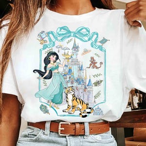 K&ouml;nnte beinhalten: Wei&szlig;es T-Shirt mit einer Cartoon-Illustration einer Prinzessin, eines Tigers und eines Schlosses. Das Design umfasst eine t&uuml;rkisfarbene Schleife und einen Rahmen mit zus&auml;tzlichen Charakteren und Elementen. Die Prinzessin tr&auml;gt ein t&uuml;rkisfarbenes Outfit und h&auml;lt eine goldene Lampe.