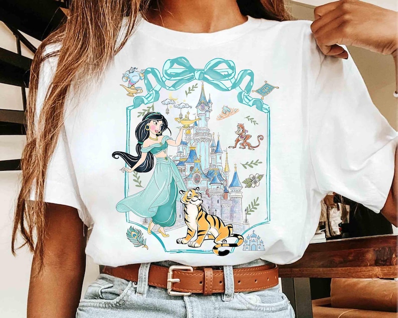 K&ouml;nnte beinhalten: Wei&szlig;es T-Shirt mit einer Cartoon-Illustration einer Prinzessin, eines Tigers und eines Schlosses. Das Design umfasst eine t&uuml;rkisfarbene Schleife und einen Rahmen mit zus&auml;tzlichen Charakteren und Elementen. Die Prinzessin tr&auml;gt ein t&uuml;rkisfarbenes Outfit und h&auml;lt eine goldene Lampe.