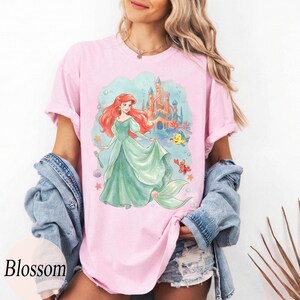 K&ouml;nnte beinhalten: Hellrosa T-Shirt mit einer Aquarellillustration einer Meerjungfrau in einem gr&uuml;nen Kleid, rotem Haar, einem Schloss und Meeresbewohnern. Das Wort "Blossom" ist unten links in Wei&szlig; aufgedruckt.