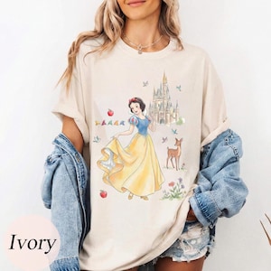 K&ouml;nnte beinhalten: Elfenbeinfarbenes T-Shirt mit einer Aquarellillustration von Schneewittchen in einem gelben Kleid, blauem Oberteil und roter Schleife. Ein Schloss, ein Reh und &Auml;pfel sind ebenfalls abgebildet. Das Shirt hat eine lockere Passform.