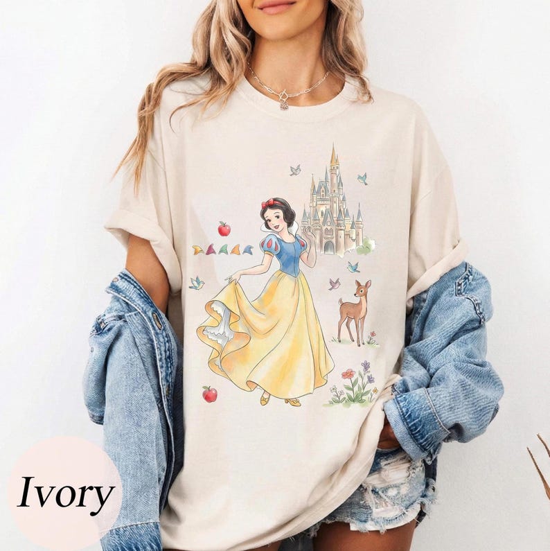 K&ouml;nnte beinhalten: Elfenbeinfarbenes T-Shirt mit einer Aquarellillustration von Schneewittchen in einem gelben Kleid, blauem Oberteil und roter Schleife. Ein Schloss, ein Reh und &Auml;pfel sind ebenfalls abgebildet. Das Shirt hat eine lockere Passform.