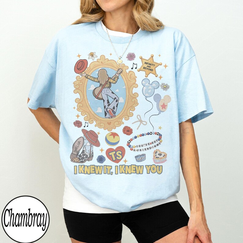 May include: A light blue t-shirt featuring a graphic print of a person with a guitar inside an ornate gold frame, surrounded by icons including a sheriff badge with the text INFINITY AND BEYOND, balloons, a cowboy boot, and the text I KNEW IT, I KNEW YOU.