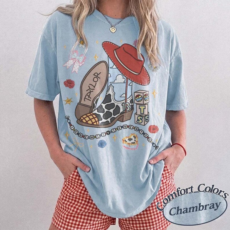 May include: A light blue t-shirt featuring a graphic print of a cow-print cowboy boot, a red cowboy hat, alphabet blocks spelling T S, a bow, and flowers. The text "TAYLOR" is on the boot, and "I KNEW YOU WERE TROUBLE" is written in a circle.
