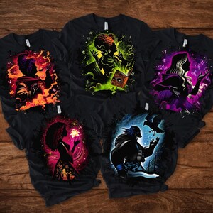 May include: Five black t-shirts with colorful graphic designs. Each shirt features a silhouette of a person with magical elements. The designs include flames, glowing books, and mystical figures. The shirts are displayed on a wooden surface.
