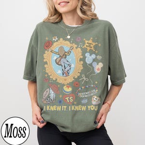 May include: An olive green t-shirt featuring a graphic print of a person playing a guitar inside an ornate gold frame, surrounded by illustrations of a cowboy hat, a star with the text INFINITY AND BEYOND, balloons, and the text I KNEW IT, I KNEW YOU.