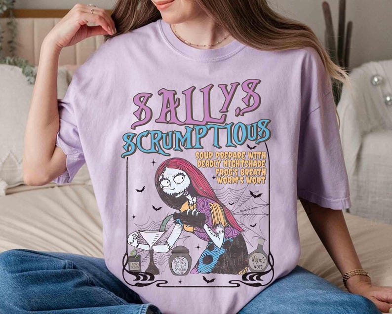 May include: A light purple t-shirt featuring a graphic of Sally from The Nightmare Before Christmas. The graphic includes the text "Sally's Scrumptious" and a recipe. The design is in shades of purple, teal, and black.