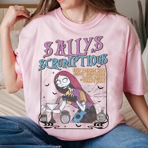 May include: Pink t-shirt featuring a graphic of Sally from The Nightmare Before Christmas. The design includes the text "Sally's Scrumptious" and a recipe. The artwork is in a vintage, cartoon style, with bats and a spiderweb.