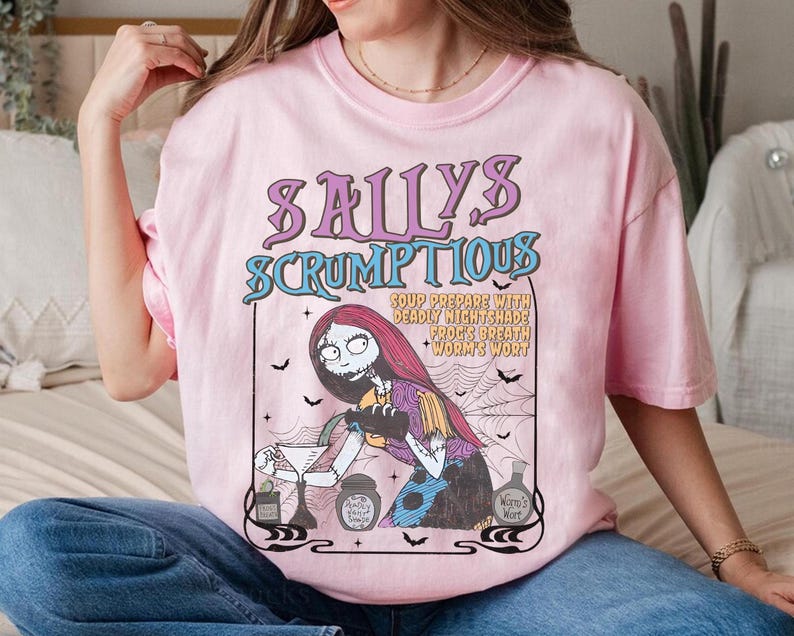 May include: Pink t-shirt featuring a graphic of Sally from The Nightmare Before Christmas. The design includes the text "Sally's Scrumptious" and a recipe. The artwork is in a vintage, cartoon style, with bats and a spiderweb.