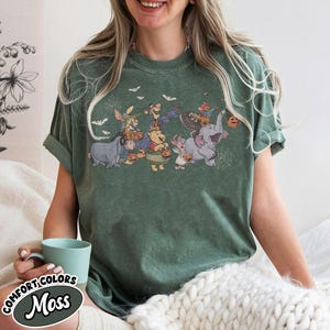 May include: Green t-shirt featuring a Halloween-themed graphic of cartoon characters, including Winnie the Pooh and friends, dressed in costumes. The shirt has rolled-up sleeves and the text "COMFORT COLORS Moss" is visible on a label.