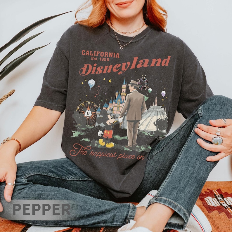 Puede incluir: Camiseta gris oscuro con un gr&aacute;fico de Disneyland. El gr&aacute;fico presenta a un hombre con traje, Mickey Mouse y el castillo de Disneyland. El texto en la camiseta dice "CALIFORNIA Est. 1955 Disneyland" y "The happiest place on".