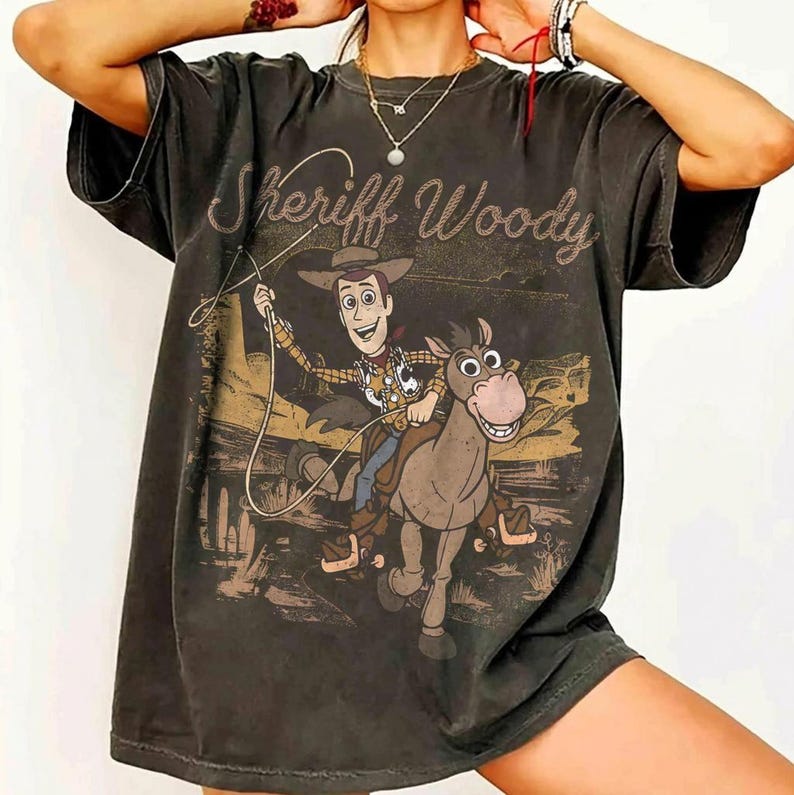 Pode incluir: Camiseta cinza escuro com um desenho do Xerife Woody de Toy Story montado a cavalo, e o texto "Sheriff Woody". O design usa tons de marrom, bege e bronzeado, dando um visual vintage.