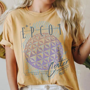 May include: Mustard yellow t-shirt featuring a graphic of the Epcot Center sphere with the text "EPCOT Center" in blue. The shirt also includes the text "Walt Disney World" and the year "1982".