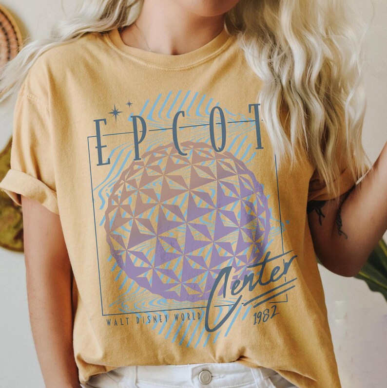 May include: Mustard yellow t-shirt featuring a graphic of the Epcot Center sphere with the text "EPCOT Center" in blue. The shirt also includes the text "Walt Disney World" and the year "1982".