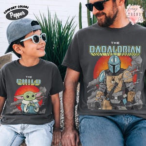 May include: Two charcoal gray t-shirts featuring Star Wars graphics. The smaller shirt displays The Child with a red sun background and text. The larger shirt features The Dadalorian with a Mandalorian character, red sun, and smoke graphics.