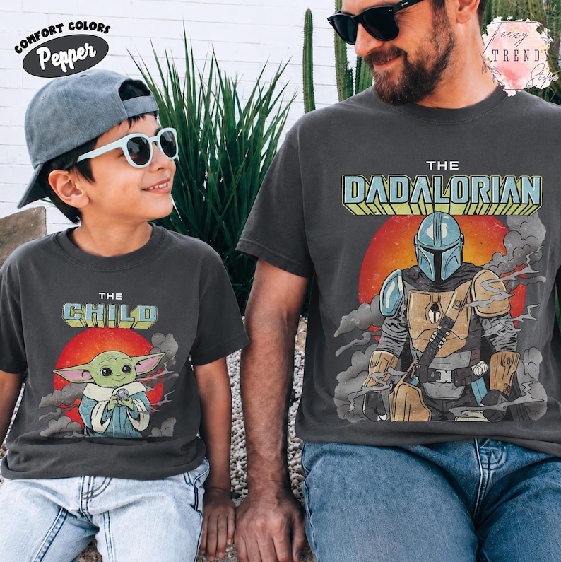May include: Two charcoal gray t-shirts featuring Star Wars graphics. The smaller shirt displays The Child with a red sun background and text. The larger shirt features The Dadalorian with a Mandalorian character, red sun, and smoke graphics.
