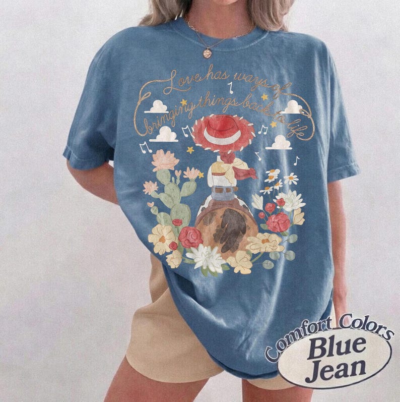 May include: A blue t-shirt featuring a graphic of a cowgirl character wearing a red hat, surrounded by cacti, flowers, clouds, and musical notes. The text reads, Love has ways of bringing things back to life. The shirt has a crew neck and short sleeves.