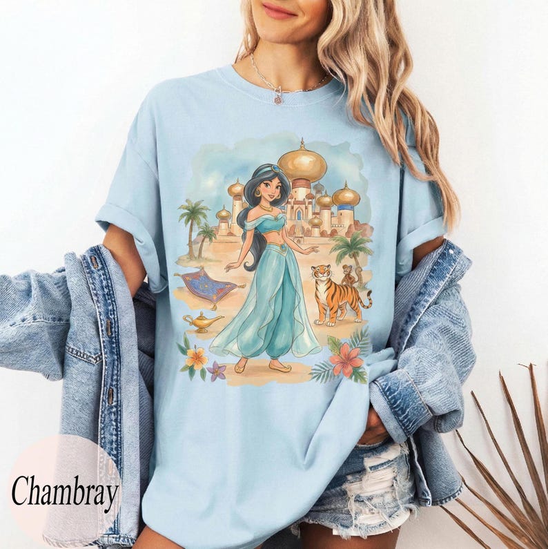 K&ouml;nnte beinhalten: Hellblaues T-Shirt mit einer Aquarellillustration einer Prinzessin in Teal vor einem Palast mit einem Tiger und Affen. Das Wort "Chambray" ist unten links aufgedruckt.
