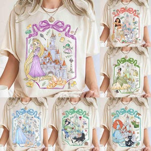 K&ouml;nnte beinhalten: Cremefarbene T-Shirts mit verschiedenen grafischen Designs zum Thema Prinzessinnen. Jedes Shirt zeigt eine andere Prinzessin, ein Schloss und verwandte Bilder in einem dekorativen Rahmen. Die Designs umfassen Rapunzel, Moana, Tiana, Elsa, Mulan und Merida.