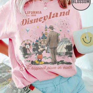 Puede incluir: Camiseta rosa de Disneyland con un gr&aacute;fico retro de Mickey Mouse, un hombre y el castillo de Disneyland. La camiseta tiene el texto "California Disneyland Est. 1955" y "The happiest place on earth."