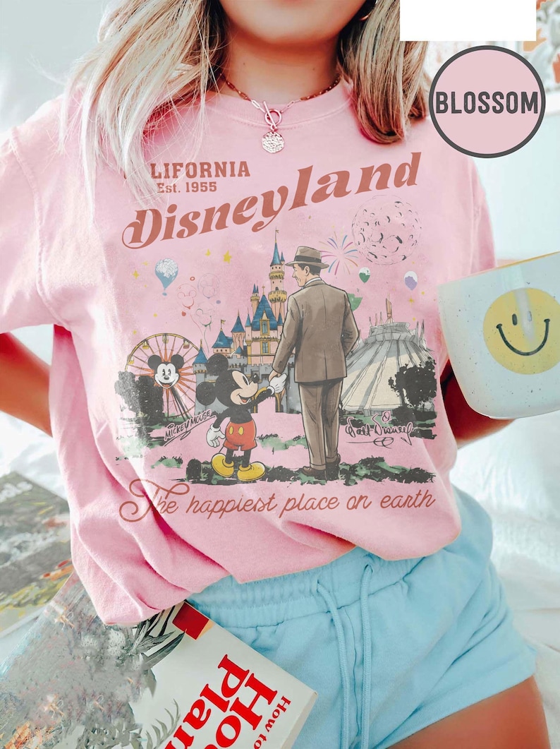Puede incluir: Camiseta rosa de Disneyland con un gr&aacute;fico retro de Mickey Mouse, un hombre y el castillo de Disneyland. La camiseta tiene el texto "California Disneyland Est. 1955" y "The happiest place on earth."