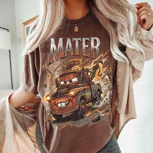 May include: Brown t-shirt featuring a graphic of Mater from the movie Cars. The design includes the word "MATER" in a distressed font above the character, with lightning bolts and tools surrounding the image. The shirt has a vintage look.