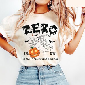 May include: Cream-colored t-shirt featuring a graphic of Zero from The Nightmare Before Christmas. The design includes the word "ZERO" in black, a pumpkin, bats, and the text "EST. 1973 THE NIGHTMARE BEFORE CHRISTMAS."
