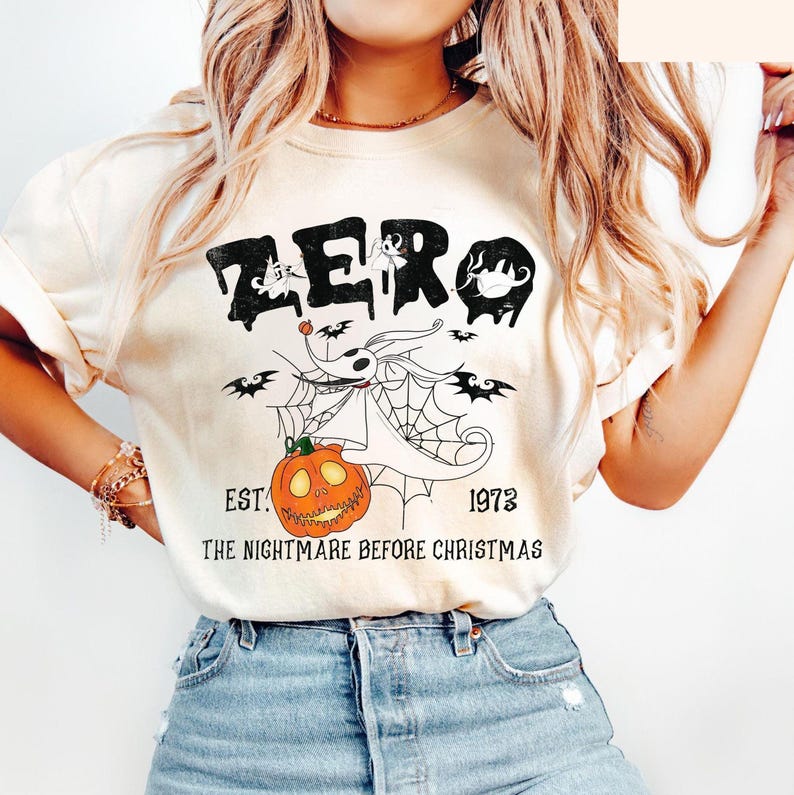 May include: Cream-colored t-shirt featuring a graphic of Zero from The Nightmare Before Christmas. The design includes the word "ZERO" in black, a pumpkin, bats, and the text "EST. 1973 THE NIGHTMARE BEFORE CHRISTMAS."
