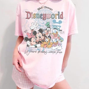 Puede incluir: Camiseta rosa claro con la inscripci&oacute;n "Walt Disney's Disneyworld" y un dibujo de Mickey, Minnie, Goofy, Donald y Daisy. La camiseta tambi&eacute;n incluye el texto "Where dreams come true" y "est. 1971".