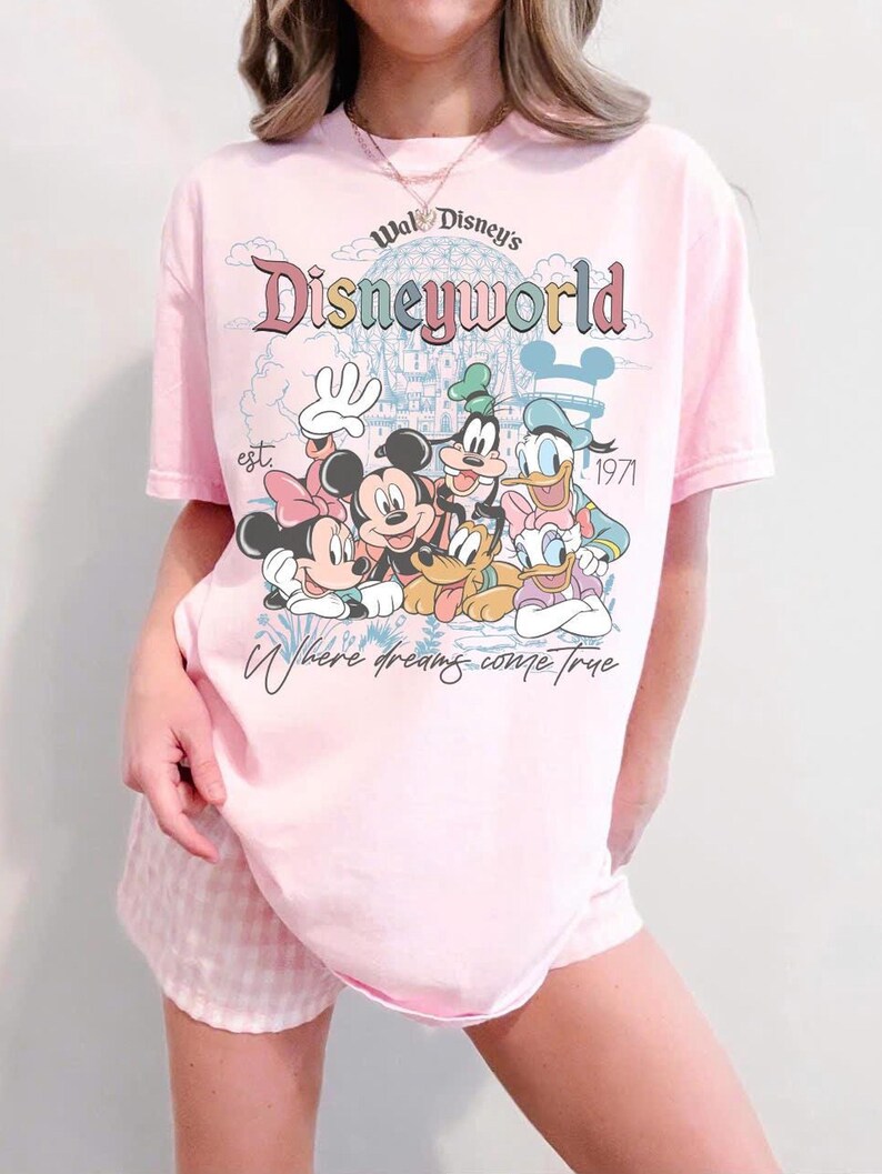 Puede incluir: Camiseta rosa claro con la inscripci&oacute;n "Walt Disney's Disneyworld" y un dibujo de Mickey, Minnie, Goofy, Donald y Daisy. La camiseta tambi&eacute;n incluye el texto "Where dreams come true" y "est. 1971".