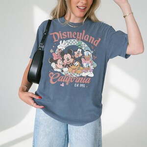 May include: Blue t-shirt featuring a Disneyland California graphic with Mickey Mouse, Minnie Mouse, Donald Duck, Goofy, and Pluto. The graphic is in pink, white, and green. The shirt has short sleeves and a crew neck.
